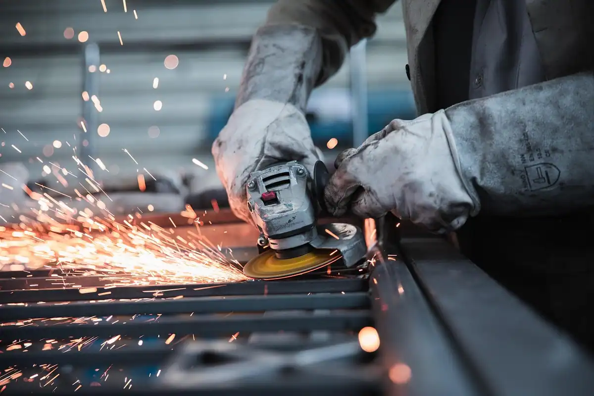 Muncitor cu mănuși care șlefuiește o piesă metalică, printre scântei, într-un atelier cu utilaje amortizabile