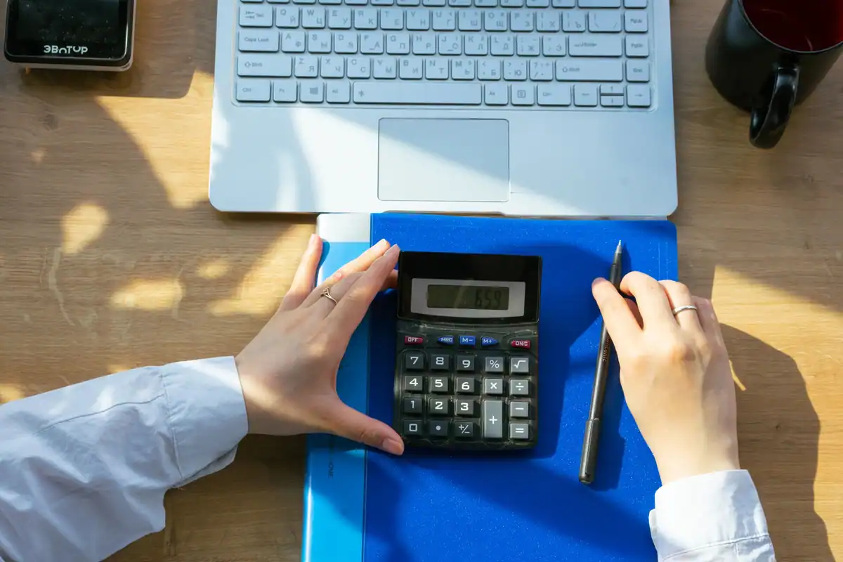 Mâini care apasă pe calculator, lângă laptop, pix și caiet, la calculul unor dividende interimare