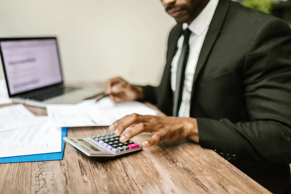 Contabil în costum care apasă pe calculator, cu laptop și documente alături, la calculul profitului