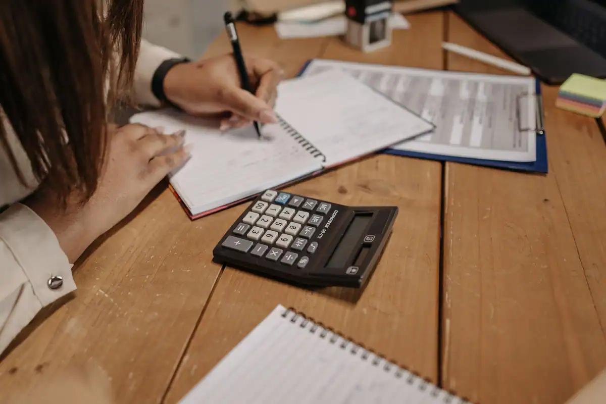 Mâini care notează într-un caiet, lângă un calculator negru, la completarea registrului de casă