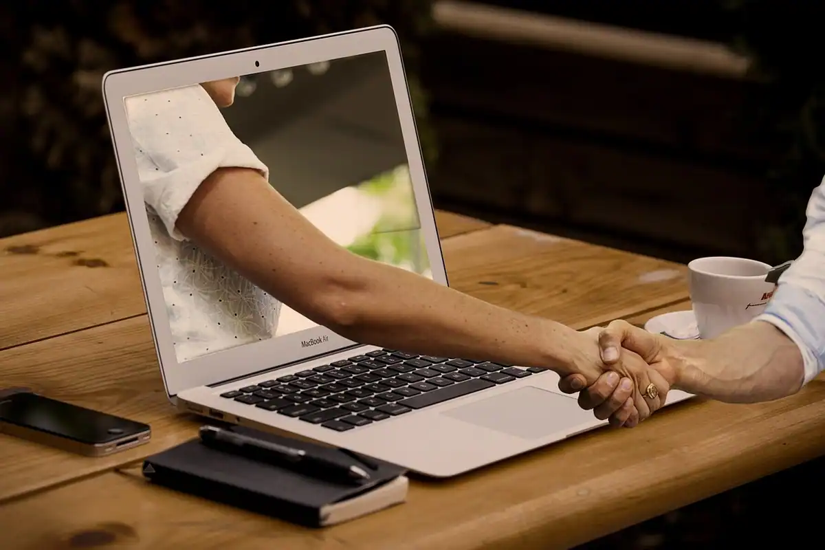 Mână care iese din ecranul unui laptop și strânge mâna unui om, simbol al serviciilor online