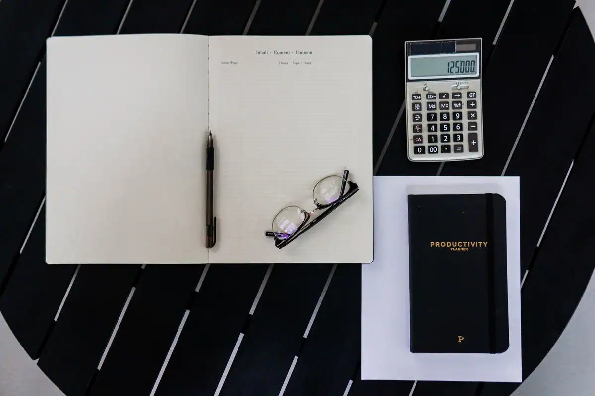 Caiet deschis, calculator, pix și ochelari pe o masă neagră, pentru calculul TVA al firmei